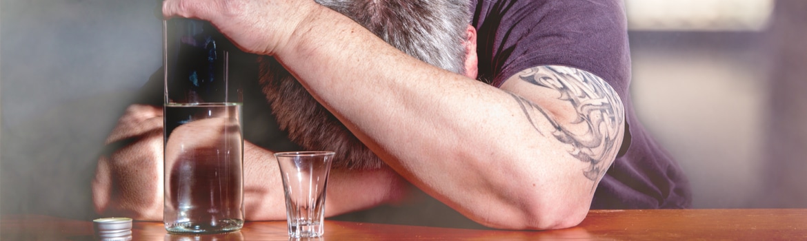 Outpatient Rehab in Colorado Pathfinders - A man sits with his head down on the bar as his left hand holds the neck of a bottle of alcohol next to his head as a shot glass sits next to him.