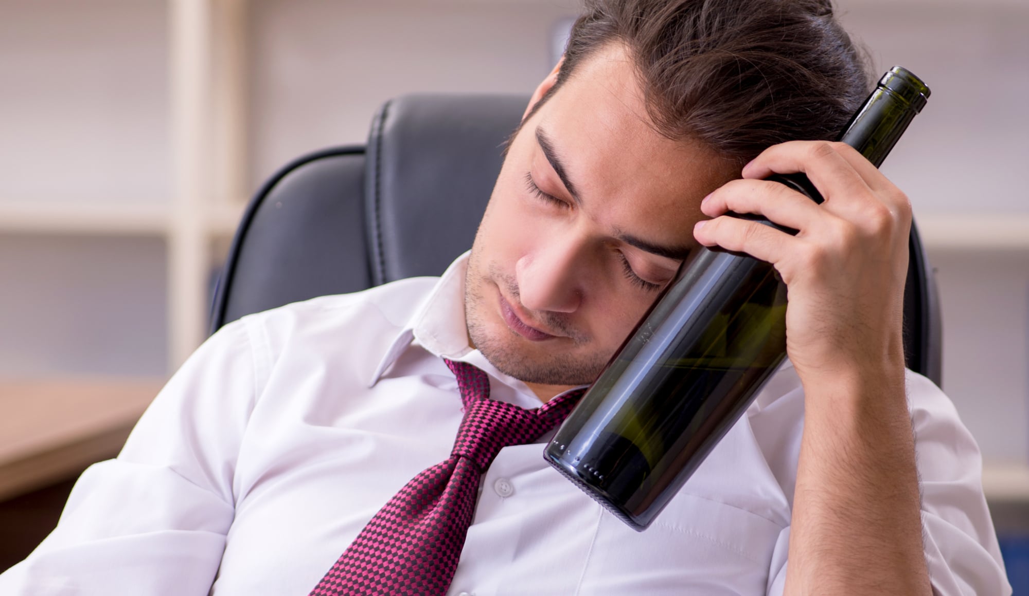 The Alarming Rates of Alcoholism Among Lawyers Pathfinders Recovery Center - A middle-aged man who works as lawyer is sitting at his desk with a bottle of alcohol as he tries to deny his alcoholism issues.