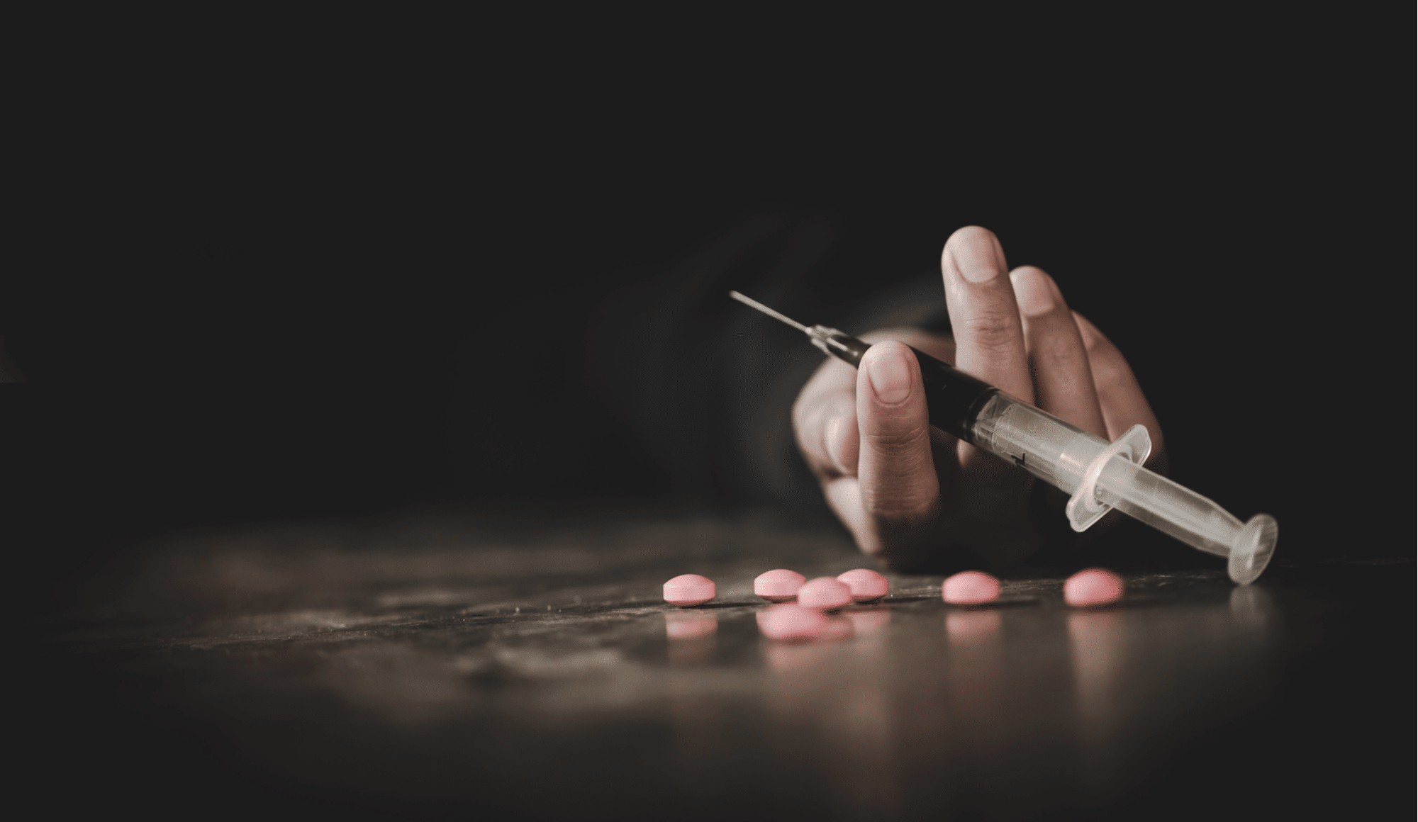 The Cost of Drugs: The Steep Price of Addiction - Pathfinders - An image of a man laying on the ground surrounded by pills and holding a needle, as he thinks about the cost of drugs in various aspects.
