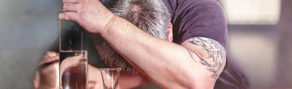  Inpatient Rehab - A close up photo of the top of a mans head as his head is down on the bar with his hand holding a bottle of alcohol.  